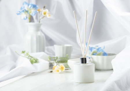 Luxury aromatic scent of reed diffuser white glass bottle is used as room freshener on the white wooden table to create relax and romantic ambient. On a background blue and white flowers.の写真素材