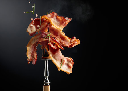 Fried bacon on a fork, sprinkled with rosemary and pink salt, against a black background with copy space.の写真素材