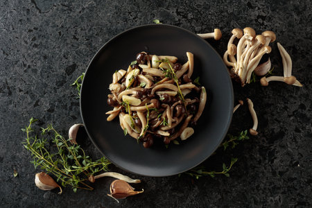 Fried shimeji mushrooms with thyme and garlic on a black plate. Fresh and prepared mushrooms with ingredients. Traditional Japanese cuisine. Top view.の写真素材