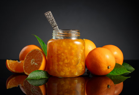 Orange jam in glass jar and fresh fruits. Sweet confiture on a black reflective background.の写真素材
