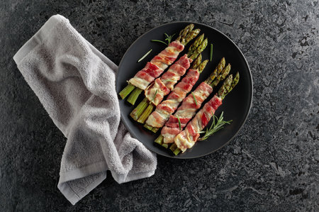 Grilled green asparagus wrapped in bacon. Homemade snack in black plate on a black stone background with copy space. Top view.の写真素材