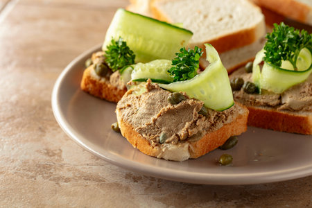 Toasts with pate on a beige plate. Open sandwiches with pate, fresh cucumber, capers, and parsley.