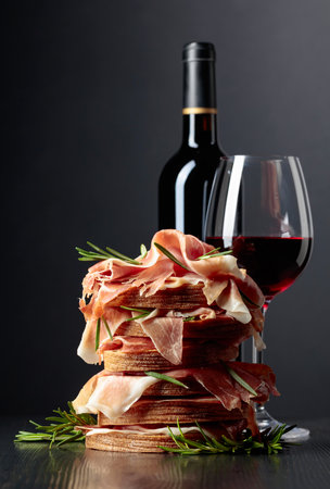 Spanish jamon or Italian prosciutto with bread, rosemary, and red wine on a black wooden table.の写真素材