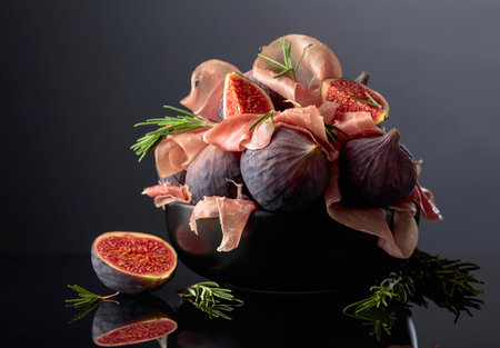 Prosciutto with rosemary and fig fruit in black dish on a black reflective background. Traditional...