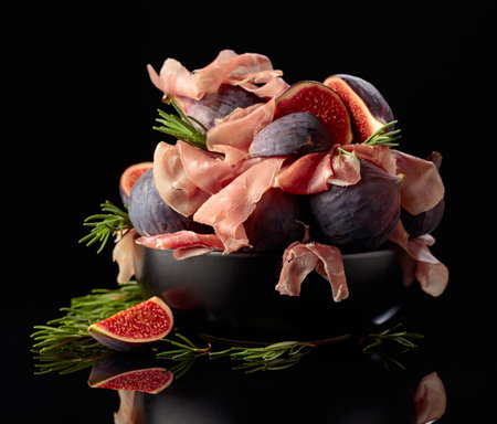 Prosciutto with rosemary and fig fruit in black dish on a black reflective background. Traditional Italian snack.の写真素材