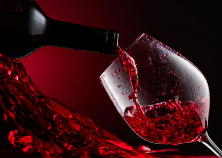 Red wine is poured from a bottle into a glass. A detailed close-up shows the rapid flow of wine from the bottle into the glass, creating natural splashes, drops, and bubbles.の写真素材