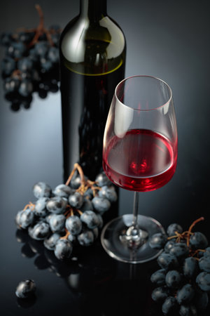 Red wine and juicy blue grapes. Bottle and glass of red wine on a black reflective background. Selective focus.の写真素材