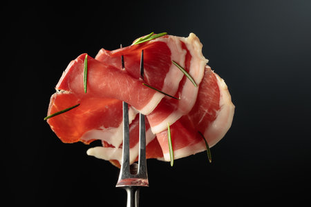 Sliced prosciutto with rosemary on a fork, black background, copy space.の写真素材