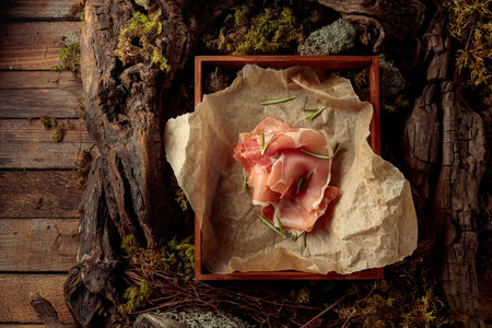 Prosciutto with rosemary on a paper in wooden box. Abstract natural composition featuring lichen, moss, and dry snags. Concept of eco products.の写真素材