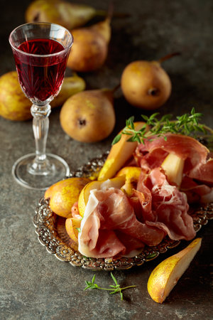 Prosciutto with pears and thyme on a silver tray. Traditional Mediterranean snack with red wine on a stone table.の写真素材