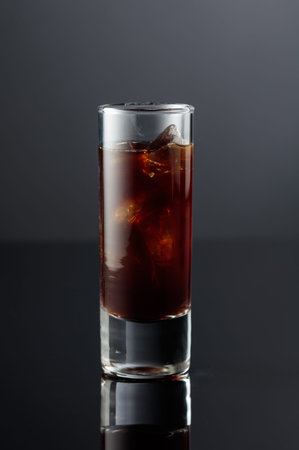 Coffee liquor with ice in tall glass on a black reflective background.