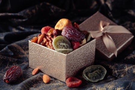 Dried tropical fruits, nuts, and raisins in gift box on a black fabric with a golden ornament.