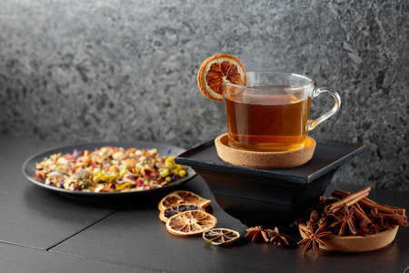 Cup of herbal tea with ingredients and slices of dried orange slices on a black ceramic table. Copy...