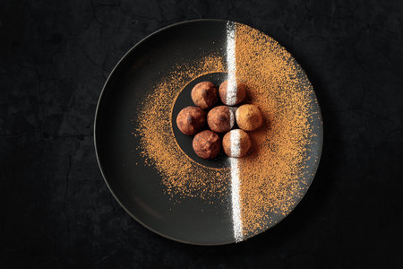 Chocolate truffles on a black plate. Sweets are sprinkled with cocoa powder. Black plate with chocolate on a black stone table. Top view. Copy space for adding text or design.