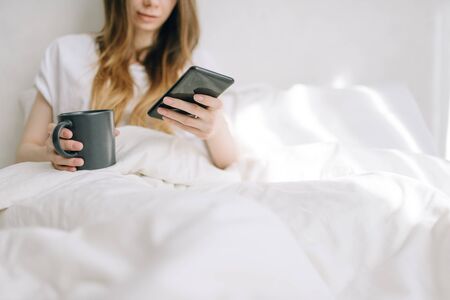 Pretty girl uses smartphone and holds cup with coffee in bed. Have a great morning at homeの写真素材
