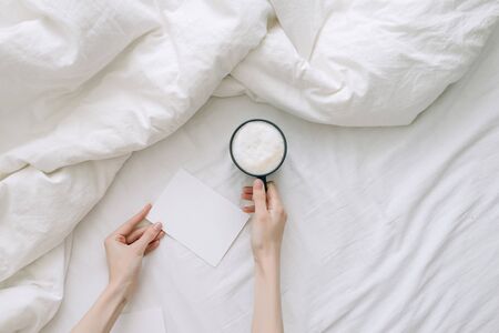 Female hand holding coffee cup and postcard in white bedclothes. Top view.の写真素材