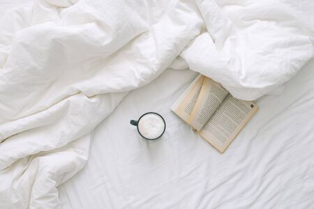 A cup of coffee and an open book lie on a white open bed. Top viewの写真素材