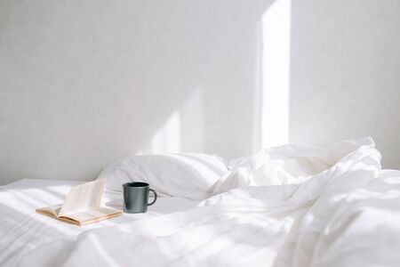 A cup of coffee and a book lying on the bed in a white bedroomの写真素材