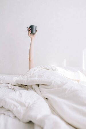A young girl holds a cup of coffee on an outstretched hand in bedの写真素材