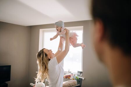 Mom holds baby in her arms in bright room at home. The husband looks at his wife and daughter as they have fun.の写真素材