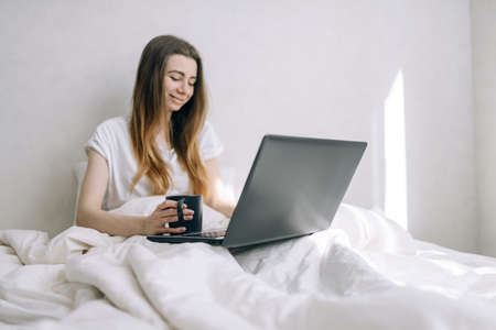 Beautiful woman in casual clothes working in the morning in bed on a laptop with a cup of coffee.の写真素材