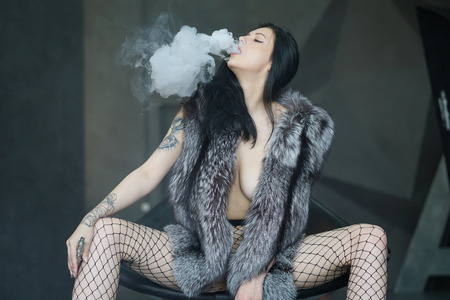 young beautiful girl posing in fur coat in studio, sitting in armchair, smokingの写真素材