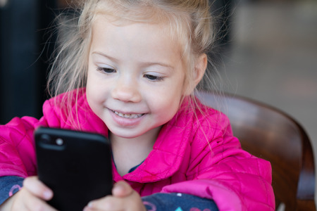 little girl is sitting with a phone in a cafeの写真素材