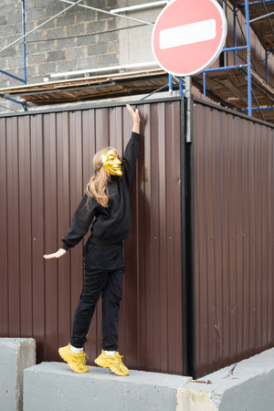 A girl in a black T-shirt and a yellow bandage on her head stands against the background of a metal fence. The concept of quarantine and self-isolation.の写真素材