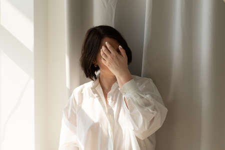 young beautiful woman posing in a white shirt in the studio, standing near the windowの写真素材