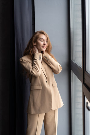 beautiful young woman in beige suit standing near window and looking awayの写真素材