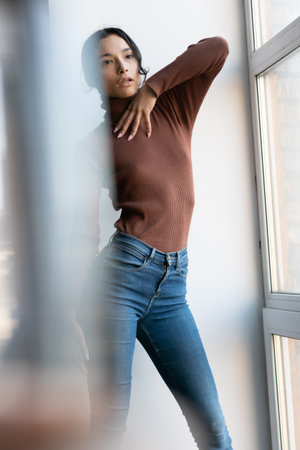 young asian woman looking at camera while standing near window at homeの写真素材