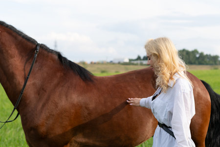 Beautiful blonde girl with long hair in a white shirt stands next to a brown horse on a green meadowの写真素材