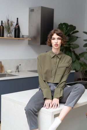 beautiful young woman sitting on table and looking at camera in kitchenの写真素材