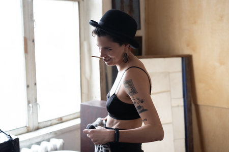 Beautiful girl with tattoos on her body in a black top and a hat posing in the interiorの写真素材