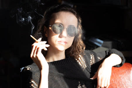 Portrait of a beautiful girl in sunglasses and a black sweater smoking a cigaretteの写真素材