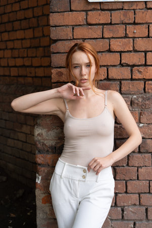 Beautiful young redhead girl in beige top and white pants posing near brick wall.の写真素材