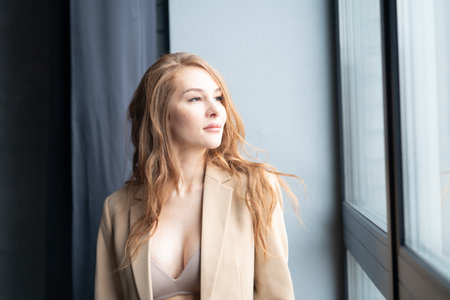 beautiful young woman in beige coat standing near window and looking awayの写真素材