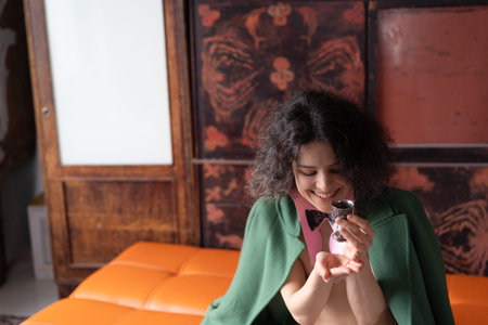 Beautiful young woman with curly hair in a green jacket sits on a orange sofa and eats chocolateの写真素材
