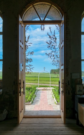 View on a rural landscape through the open doors of the Orthodox church. Passage of the temple through the iron arch with white Orthodox cross on it.の写真素材