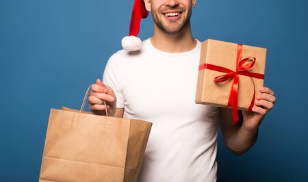 cropped view of smiling young man in santa hat holding paper bag and gift isolated on blueの写真素材
