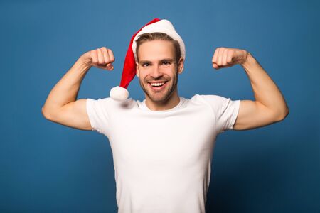 smiling young man in santa hat showing muscles isolatedの写真素材