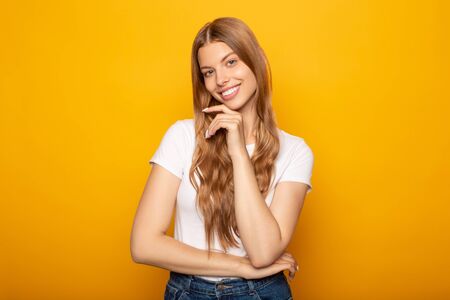 smiling young blonde girl with long hair touching chin isolated on yellow backgroundの写真素材
