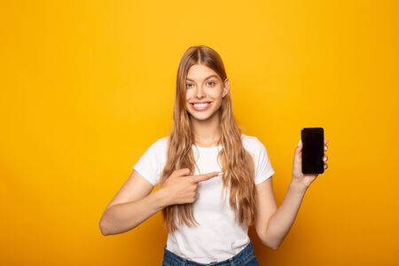 smiling girl pointing with finger at smartphone isolated on yellow backgroundの写真素材