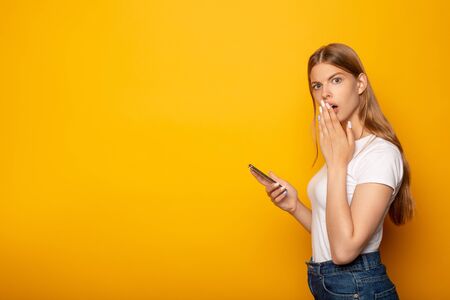 side view of shocked girl covering mouth with hand while holding smartphone isolated on yellowの写真素材