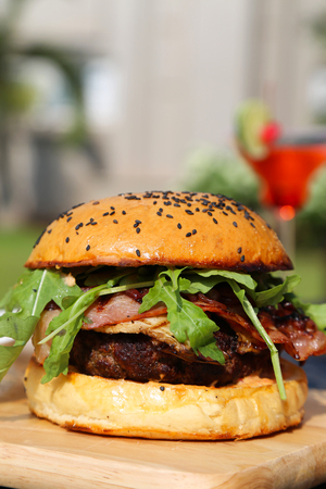 Giant Size White Bread Beef Burger with Herbs and Bacon served on a Light Color Wooden Board with a Blurred Pink Cocktail Drink in the Backgroundの写真素材