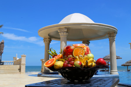 Large Plate of Oranges Bananas Apples Mango Grapes Dragon Fruits and Peaches atop Black Metal Ornamented Table with Blue Sky in the Backgroundの写真素材