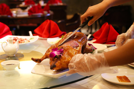 Hands of Two Waiters Cutting The Peking Duck with a Knife Decorated with Wild Purple Orchid Served on a White Plate at a Chinese Restaurant with Red Napkins China Ware and a Table in the Dark Blurred Backgroundの写真素材