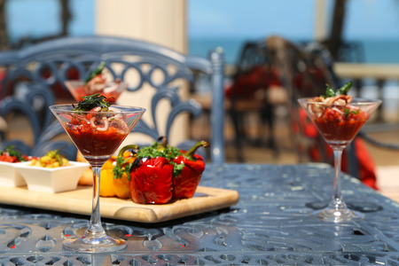 White Octopus with Tomato Mash in a Martini Glass with Peppers on a Light Wooden Board atop Black Metal Ornamented Table and Chairs with Red Pillows in the Backgroundの写真素材