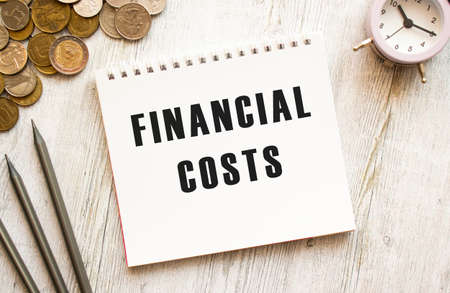 FINANCIAL COSTS text on a sheet of notepad.Coins are scattered, pencils on a gray wooden background.
