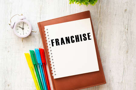 Brown notepad, inscription FRANCHISE, colored pens, clock on a wooden background.